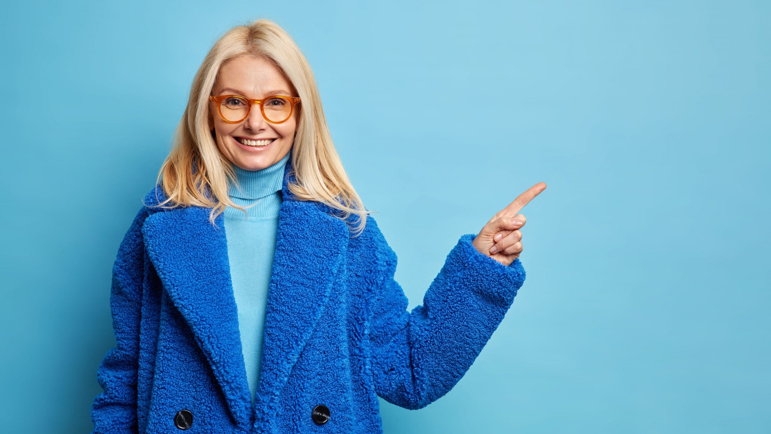 Beautiful middle aged woman with blonde hair wears spectacles and warm blue coat indicates away on copy space shows advertising content poses indoor. Winter fashion concept.